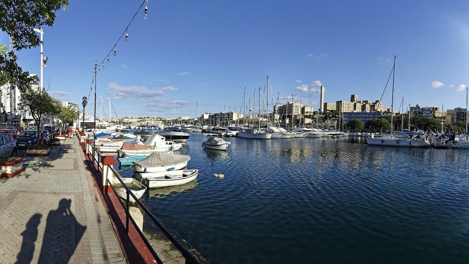 Msida Creek at the head of Marsamxett Harbour, moored yachts along both sides and small open boats drawn up by the waterfront promenade.